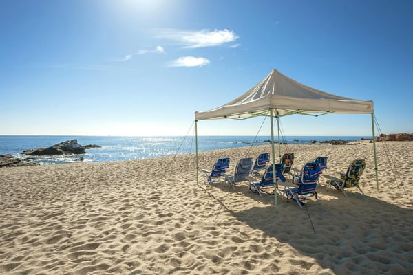 Sandy beach with tan canopy tent and blue beach chairs overlooking calm ocean and rocky coastline