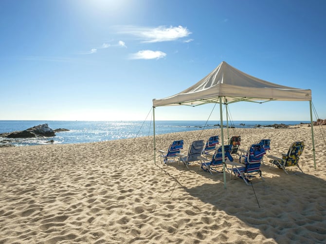 Sandy beach with tan canopy tent sheltering blue lounge chairs, ocean and rocky coastline in background