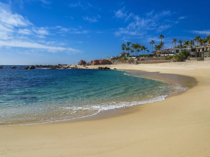 Sandy beach with turquoise water, rocky outcrops, and beachfront homes with palm trees under blue sky